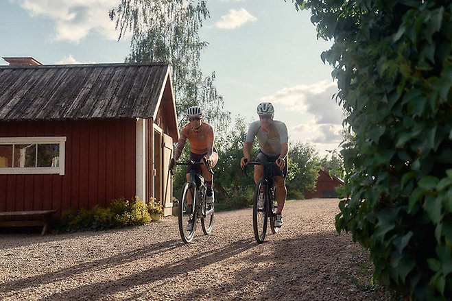 Två cyklister som cyklar på en grusväg och har precis passerat en röd byggnad.